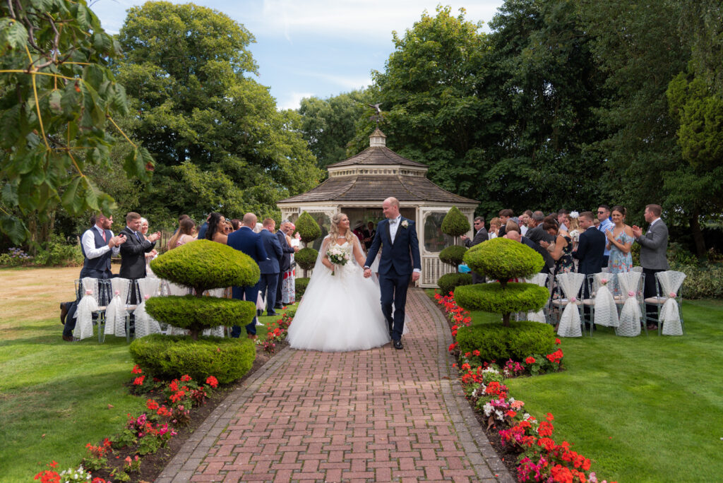 Bride and groom walk down garden aisle after an outdoor wedding at Thornton Hall