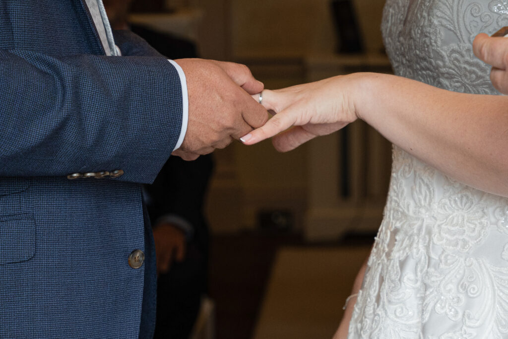 A person in a blue suit places a ring on the finger of another person wearing a white lace wedding dress.