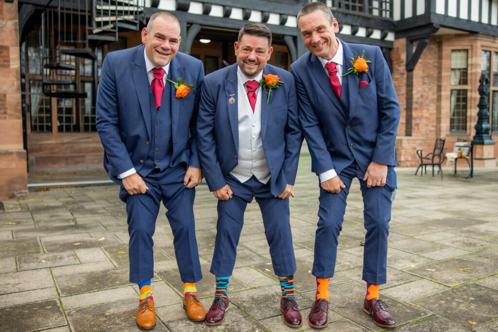 Three men in blue suits, white shirts, and red ties stand outside a building, smiling and displaying their colorful, mismatched socks.