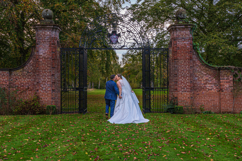 A bride and groom in wedding attire kiss in front of an ornate wrought iron gate and brick wall in a park-like setting.