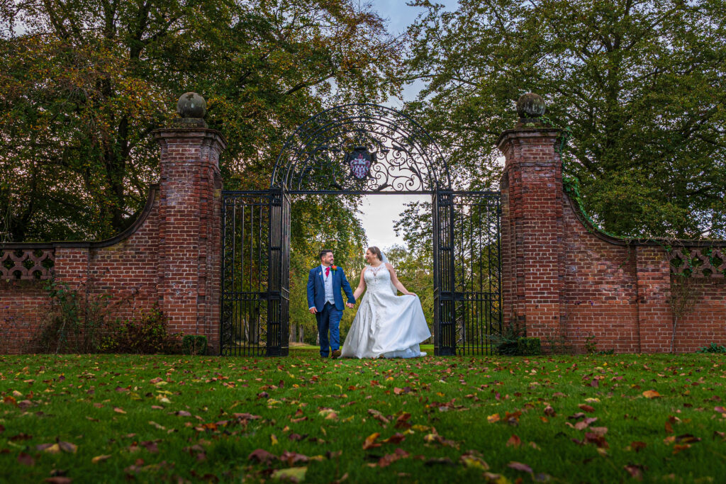 A bride and groom walk hand in hand on a grassy lawn in front of an ornate iron gate and brick wall, surrounded by trees with scattered autumn leaves.