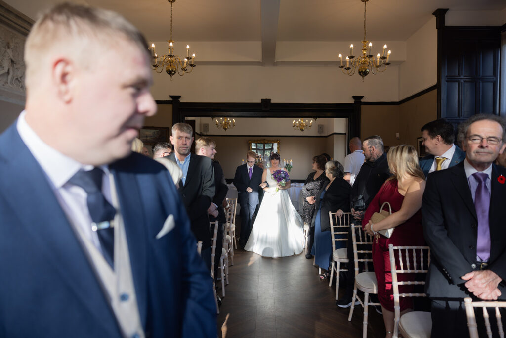 A bride and groom walk down the aisle together in a sunlit room filled with seated and standing guests at their Walton Hall Wedding Photography ceremony.