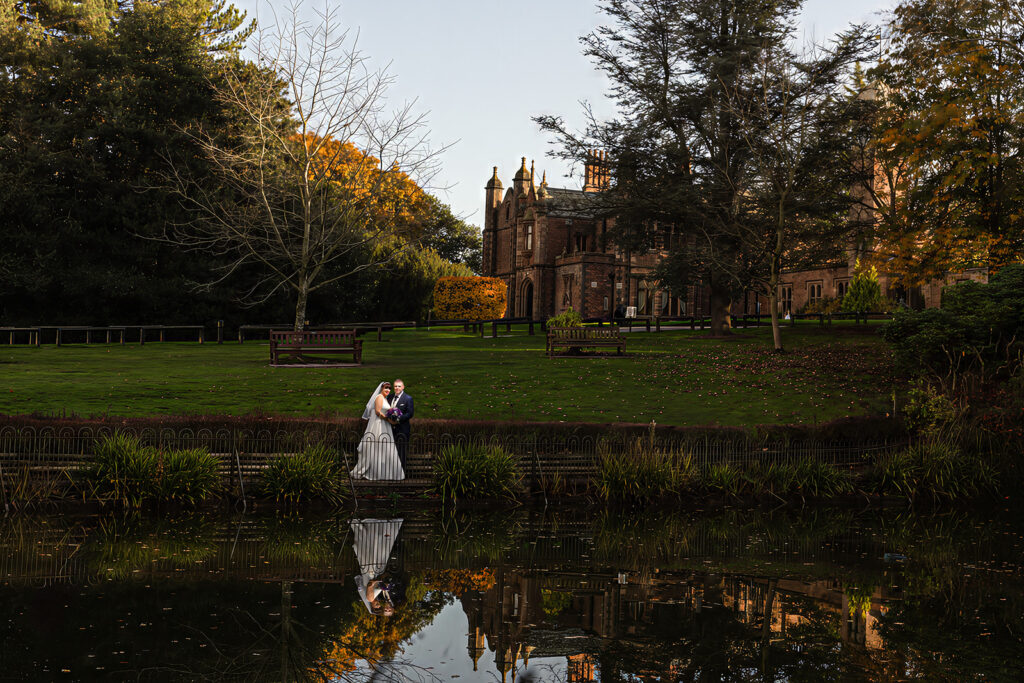 A bride and groom stand together by the pond, at Walton Hall Wedding Venue. Their reflection shimmering in the water, with a grand brick building and trees behind them—capturing the timeless elegance of Walton Hall Wedding Photography.