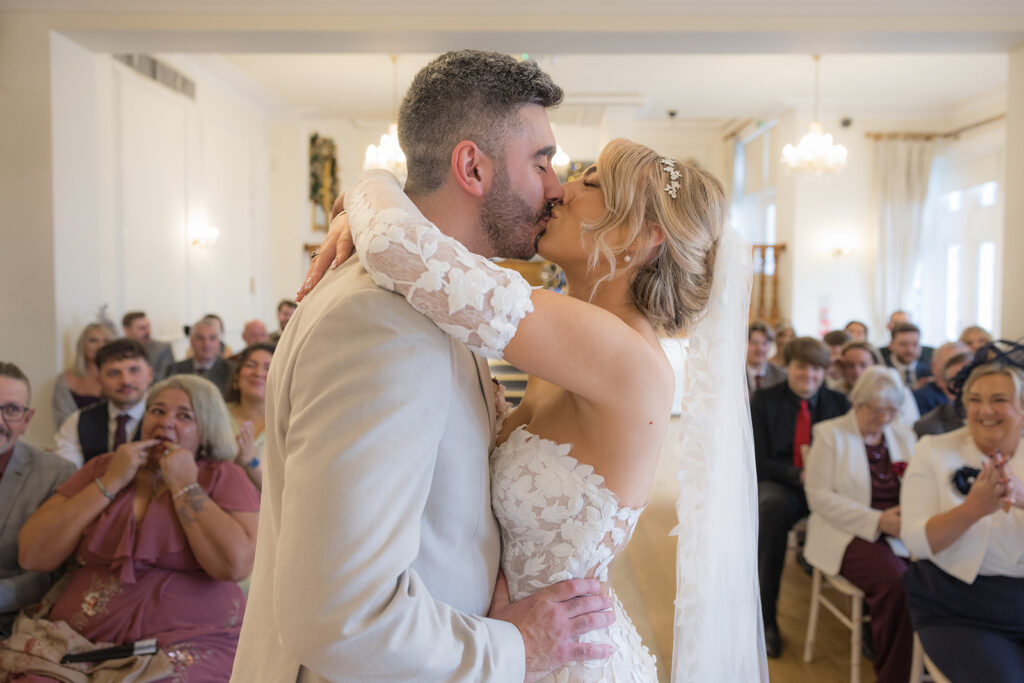 A bride and groom kiss at their wedding ceremony while guests watch and smile in a bright, elegantly decorated Lancashire Wedding Venue.