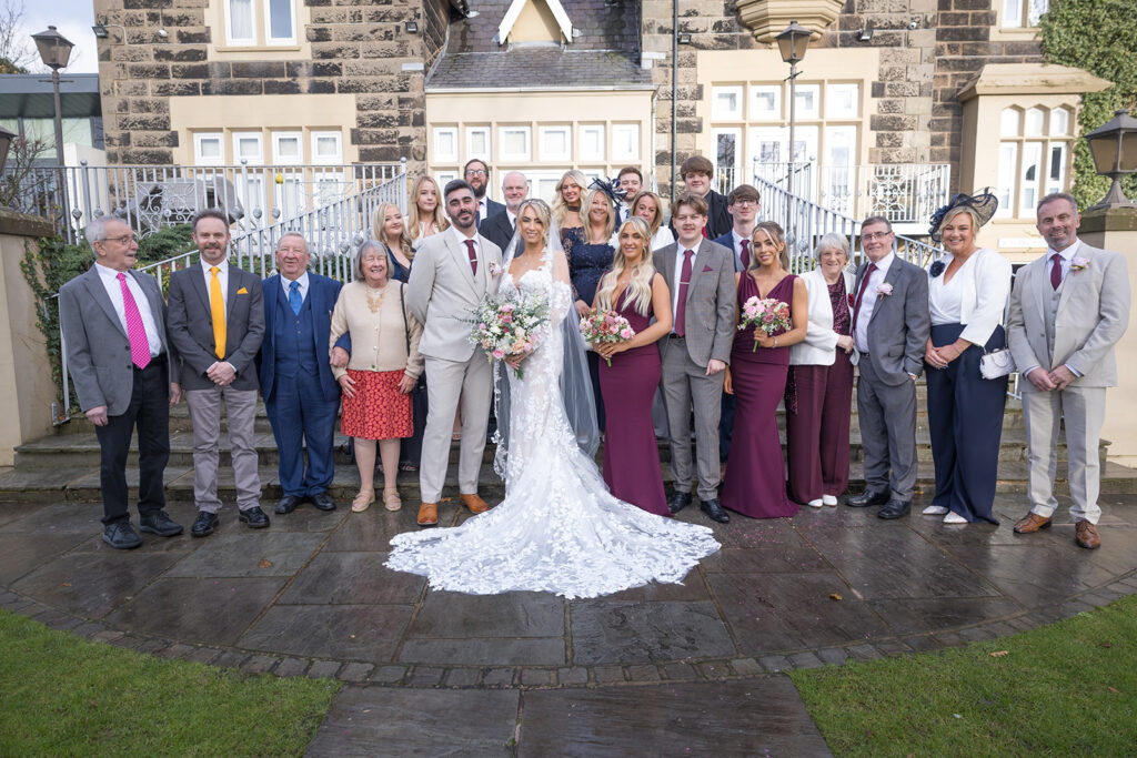 A wedding party poses for a formal group photo at West Tower Wedding, with the bride and groom in the centre surrounded by family and friends outside a beautiful stone building.