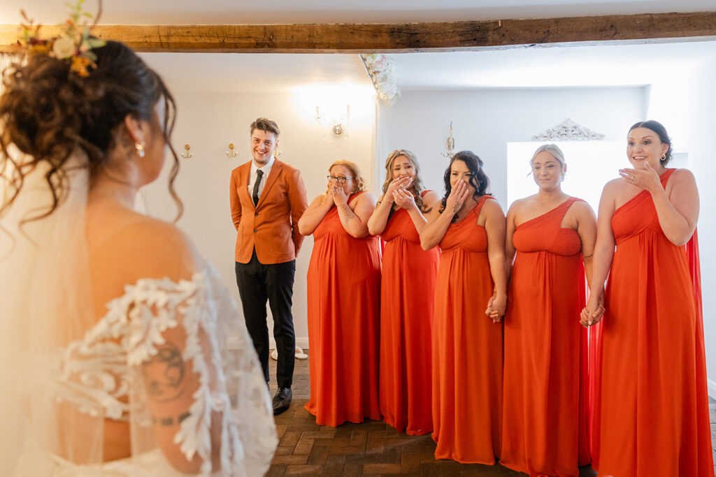 Bride reveals to excited bridesmaids in orange dresses. Alcumlow Wedding Barn Photography