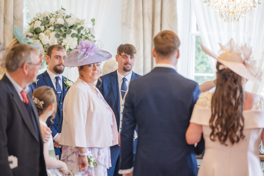 A group of people dressed formally are gathered indoors at a wedding ceremony, with floral arrangements and light streaming through large windows.