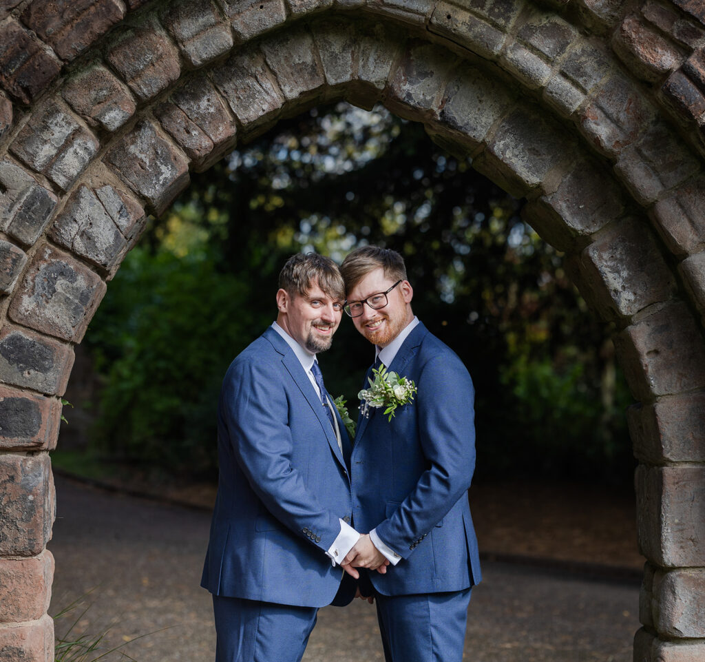 Two grooms holding hands under stone archway.