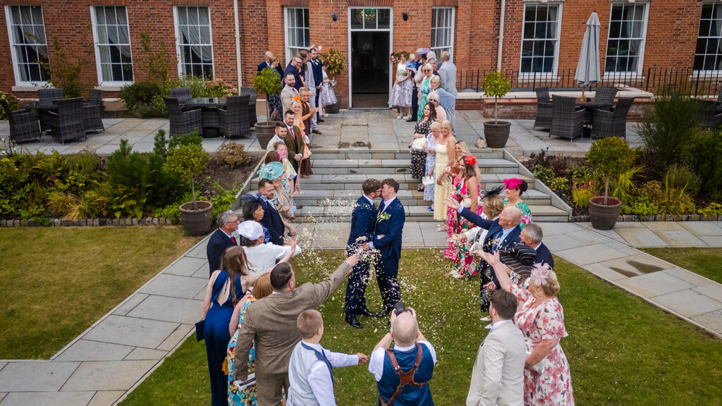 Two grooms kiss while wedding guests stand in lines on either side, throwing confetti at them outside a brick building.