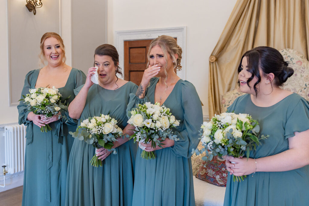 Four bridesmaids in matching green dresses stand indoors holding bouquets, reacting emotionally, with some wiping tears or covering their mouths.