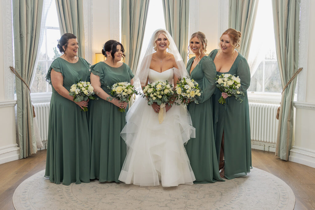 A bride in a white gown and veil stands with four bridesmaids in matching green dresses, all holding bouquets, in a bright room with large windows and curtains.