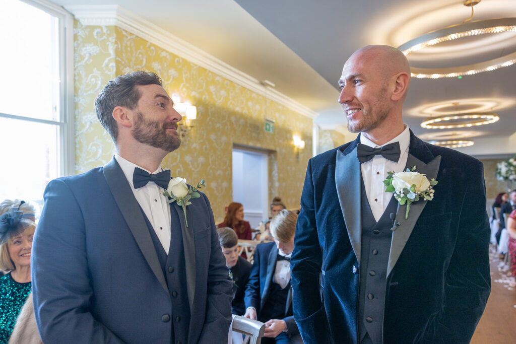 Two men in tuxedos with white boutonnieres stand indoors, facing each other and smiling, in a room decorated for a formal event.