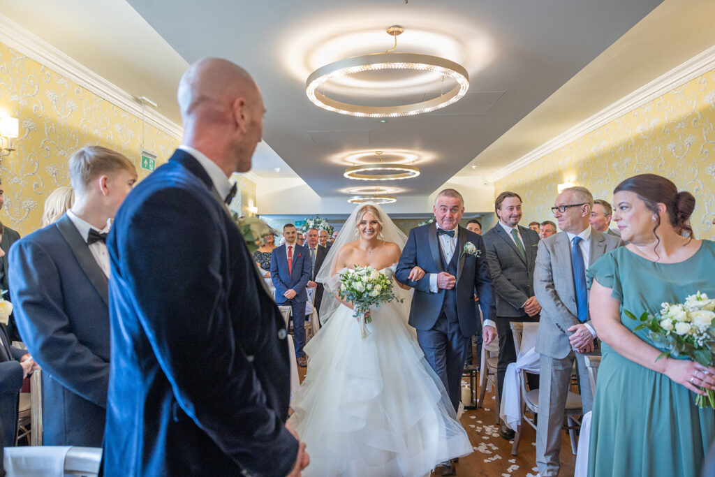 A bride in a white gown walks down the aisle with an older man, while guests on both sides watch and smile in a decorated indoor venue captured beautifully by Philip Antrobus Photography.