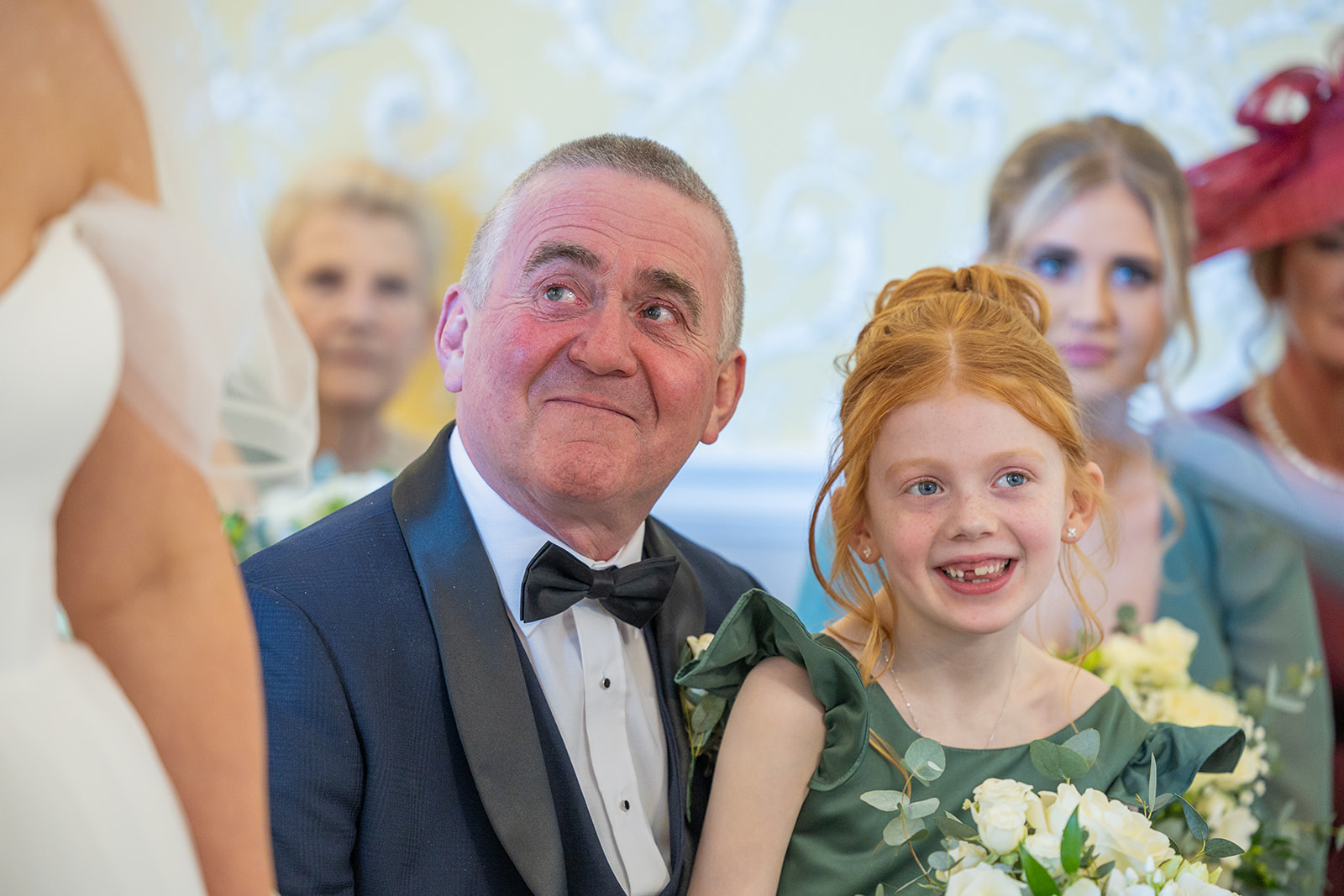 A man in a tuxedo sits next to a young girl with red hair holding flowers. Both are smiling and looking up at a bride in a white dress, capturing a joyful moment perfect for Chester wedding photography.