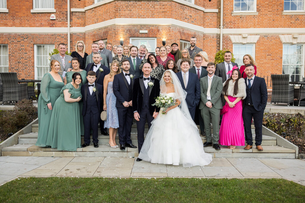 A bride and groom stand at the center of a group of wedding guests posing for a photo outside the historic Old Palace Chester.