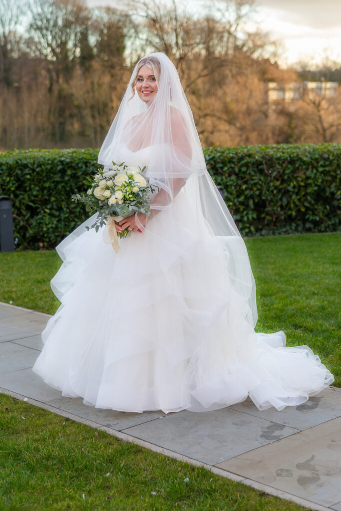 A bride in a white wedding dress and veil holds a bouquet of white flowers while standing on a stone path in the garden at an Old Palace Chester wedding.