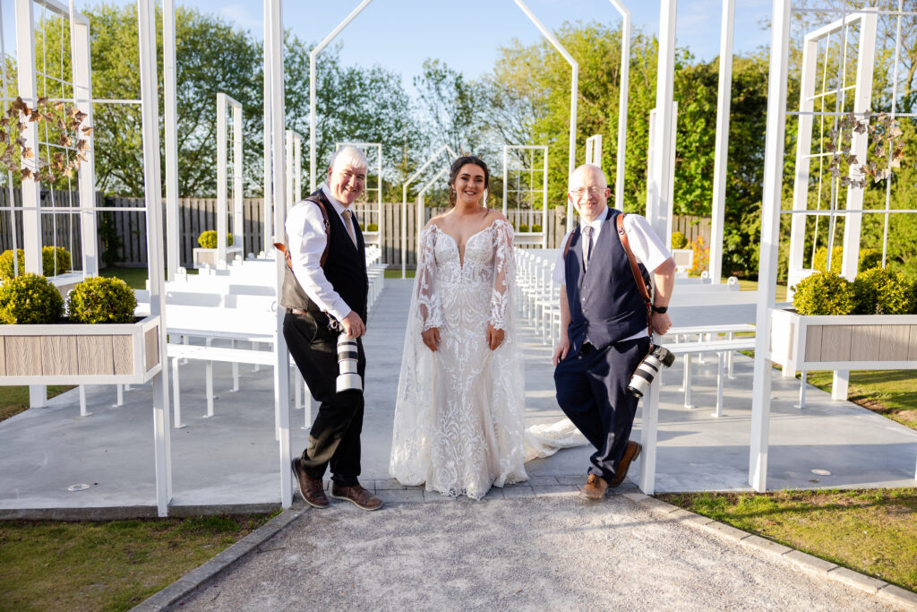 A bride in a white gown stands between two photographers holding cameras in an outdoor wedding venue with white open structures and greenery.