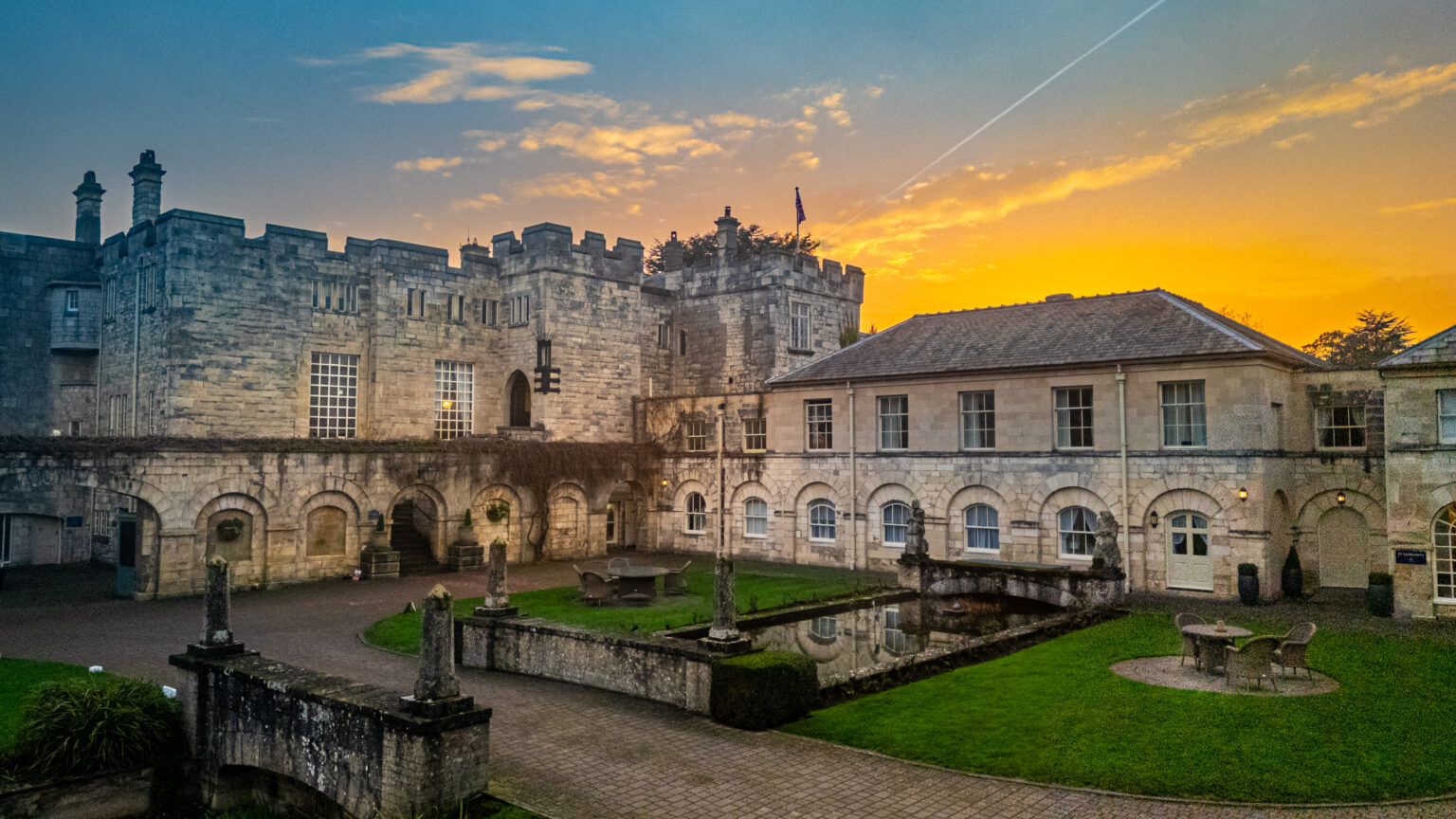 Historic stone castle and manor house with arched windows, courtyard, and small pond at sunset under an orange sky—an enchanting Hazelwood Castle Wedding Venue in Yorkshire.