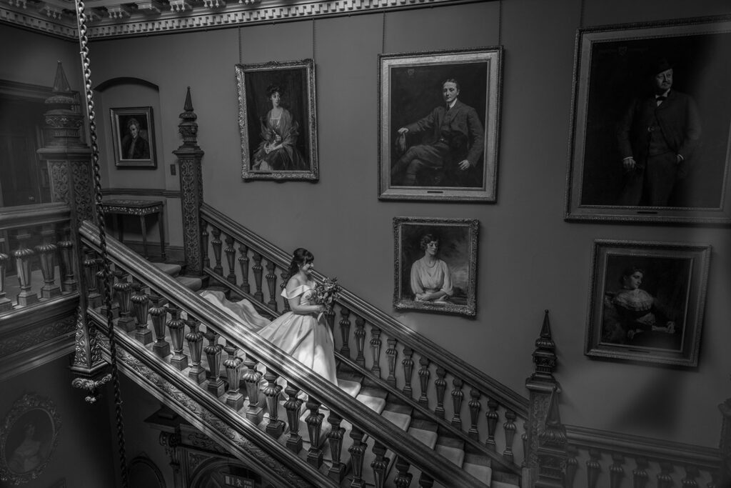 Bride descends ornate staircase surrounded by portraits.