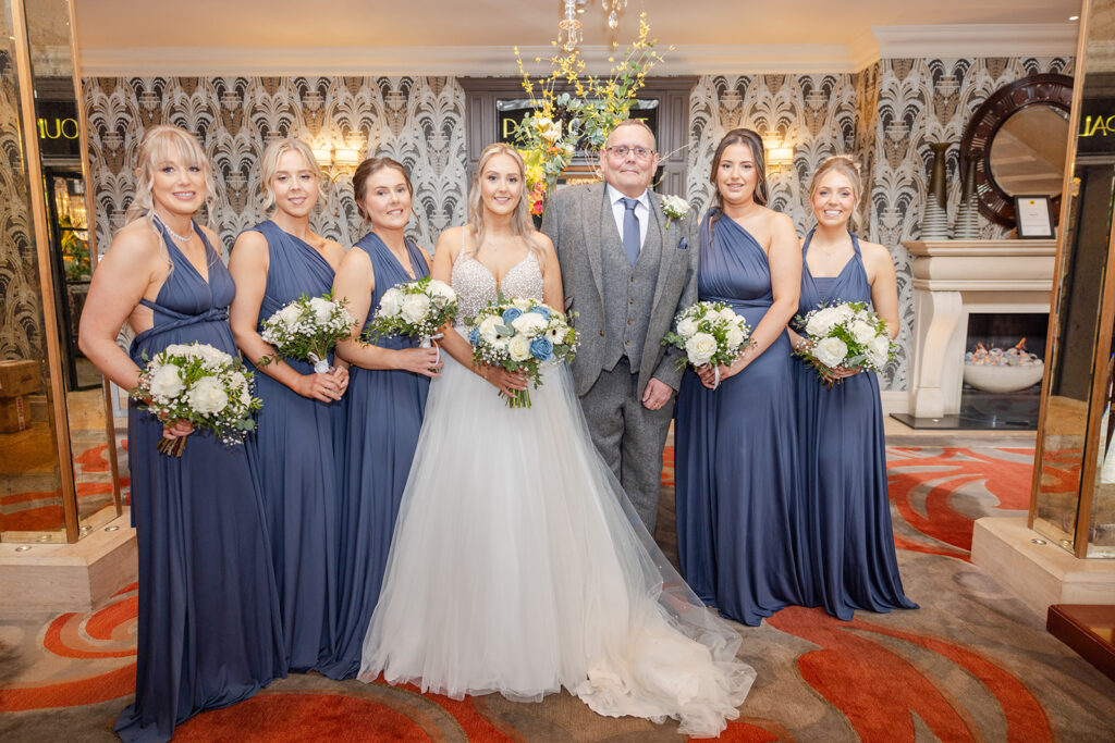 A bride in a white dress stands with six bridesmaids in blue dresses and a man in a grey suit, all holding bouquets, posing indoors at a wedding venue—captured beautifully by Philip Antrobus Photography.