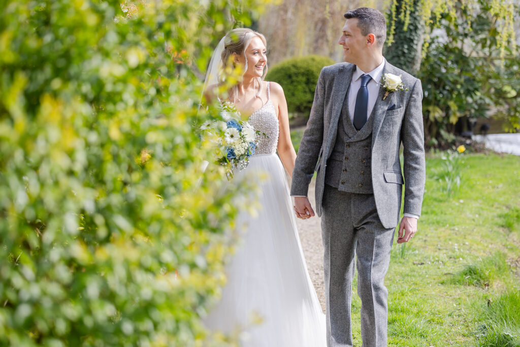 A bride in a white dress and a groom in a grey suit walk hand in hand outdoors, smiling at each other with greenery in the background, beautifully captured by Philip Antrobus Photography.