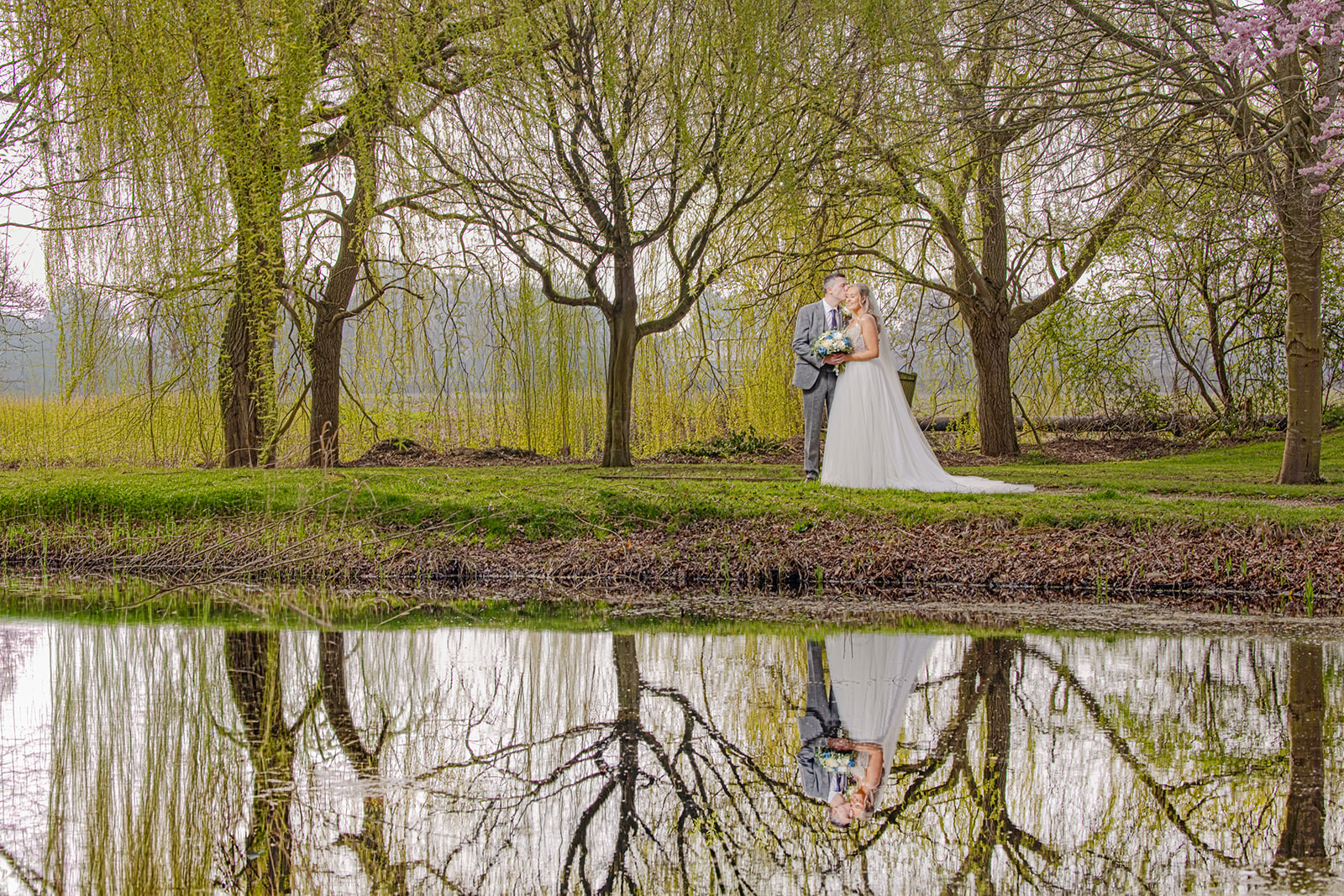 A bride and groom stand together by a pond, surrounded by trees, with their reflection visible in the water.