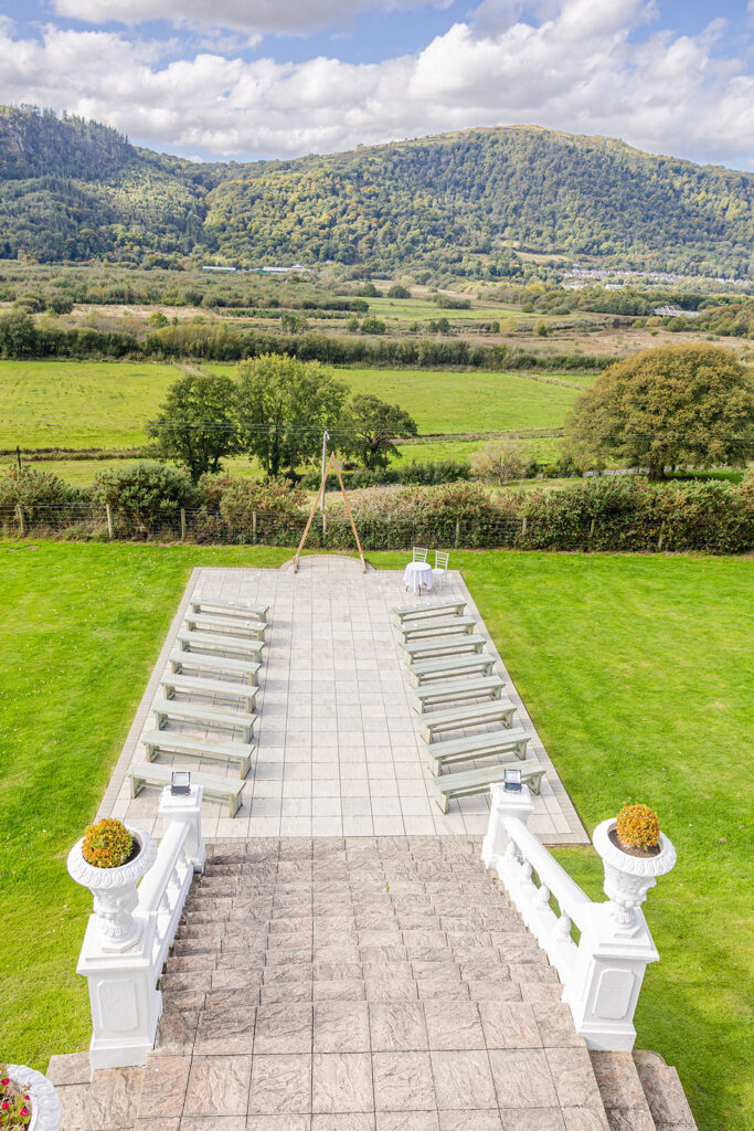 Outdoor ceremony set-up with rows of benches facing a wooden arch, surrounded by green lawns and overlooking hills and trees under a partly cloudy sky.