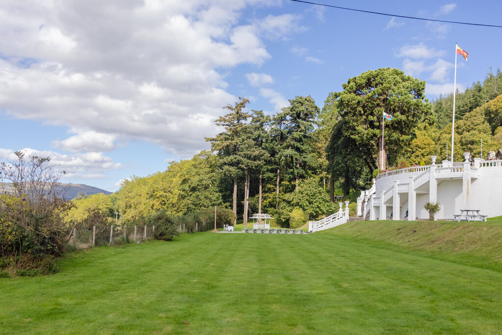 A grassy lawn leads to a white building with a staircase and flagpole, surrounded by trees and under a partly cloudy sky.