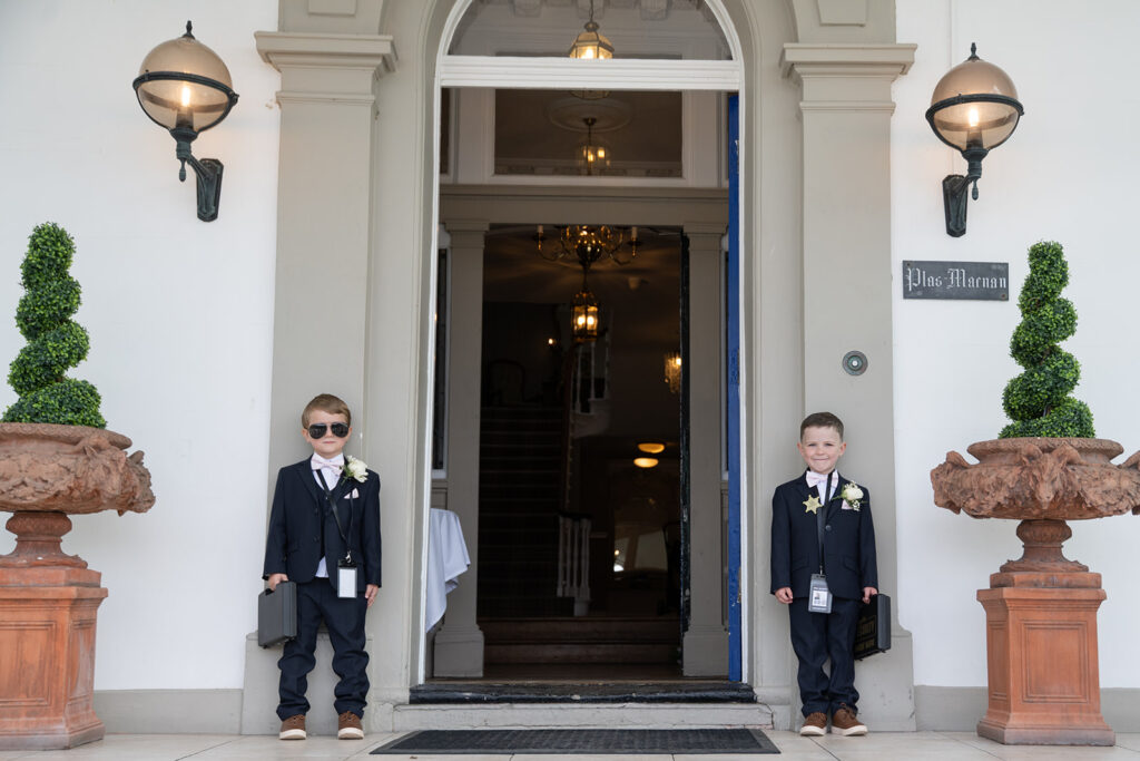 Two young boys in suits stand on either side of a building entrance, one wearing sunglasses. The entrance is flanked by potted plants and features a sign reading "Plus Merem.