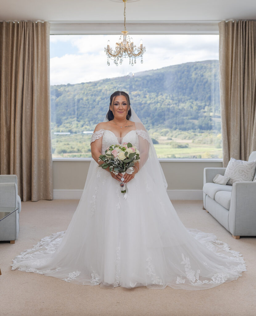 A bride in a white wedding dress holds a bouquet, standing in a bright room with large windows and mountain views in the background.