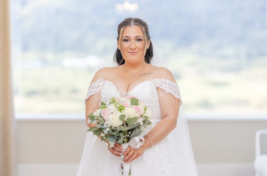 A bride in a white wedding dress holding a bunch of flowers stands indoors with a blurred outdoor background.