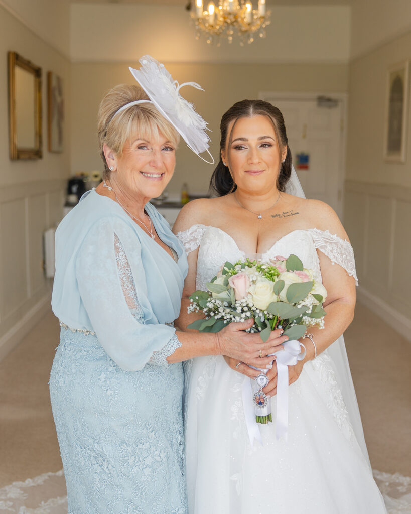 Two women stand together indoors; one in a light blue dress and hat, the other in a white wedding dress holding a bouquet. Both are smiling at the camera.