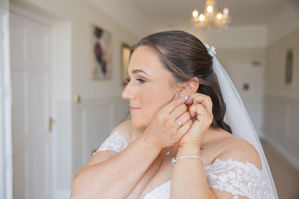 A bride with brown hair in an off-the-shoulder lace wedding dress puts on an earring in a bright room with a chandelier.