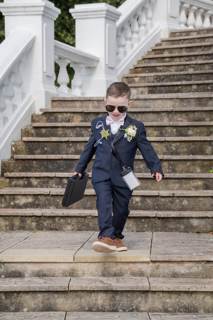 A young boy in a navy suit, sunglasses, and brown shoes walks down outdoor stone steps, holding a black briefcase, with a buttonhole and badge on his jacket.