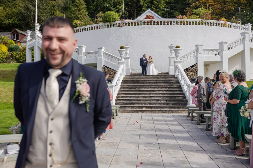 A groom stands in the foreground, smiling, whilst a bride and another person walk down the steps behind him at an outdoor wedding ceremony. Guests are seated on either side of the aisle.