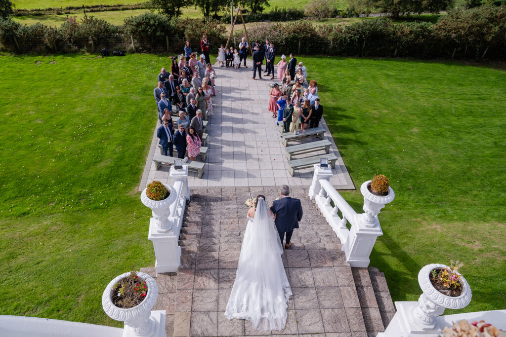 A bride walks down an outdoor aisle with a companion towards a seated group of wedding guests on a sunny day, viewed from above.