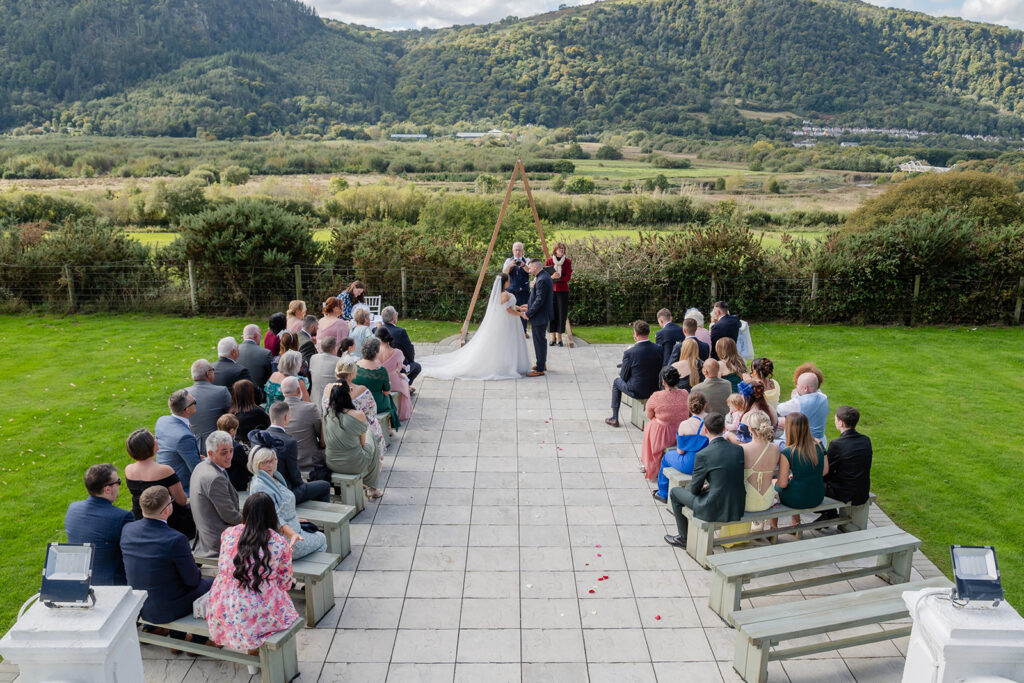 Outdoor wedding ceremony with a couple standing before an officiant, guests seated on benches, and mountains and greenery in the background.