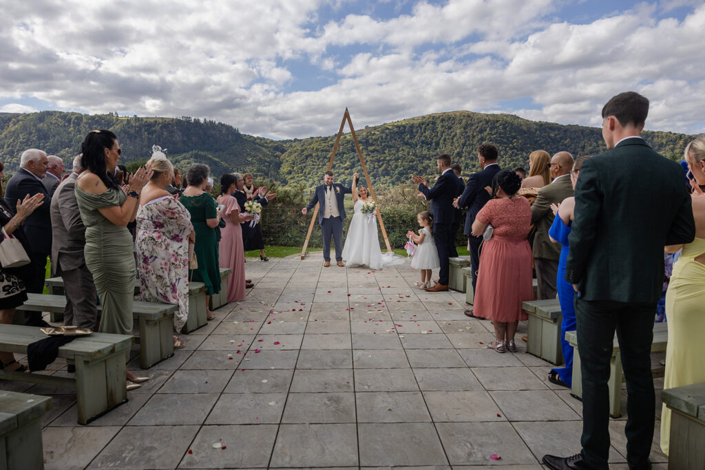 A bride and groom stand at an outdoor altar, holding hands, surrounded by wedding guests seated and standing on both sides, with mountains in the background.