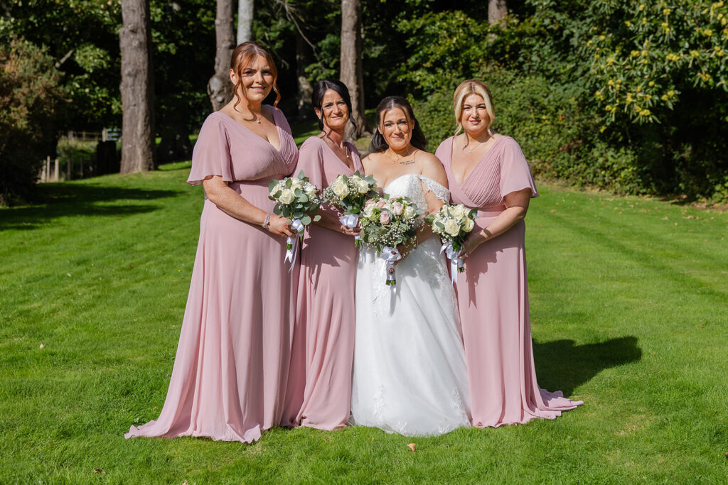 Four women posing on grass; three in matching light pink dresses hold bouquets beside one in a white wedding dress, also holding a bouquet, with trees and greenery in the background.