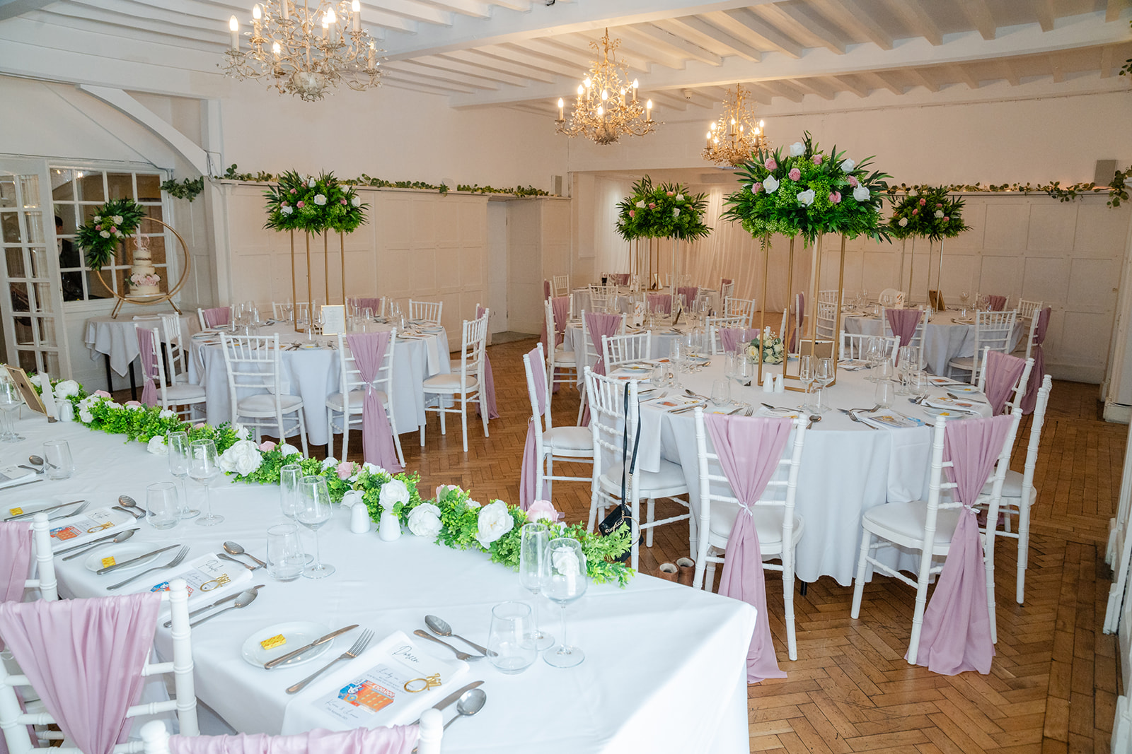 Elegant banqueting hall at Plas Maenan set for an event with round tables, white tablecloths, mauve chair sashes, floral centrepieces, and chandeliers hanging from the ceiling.