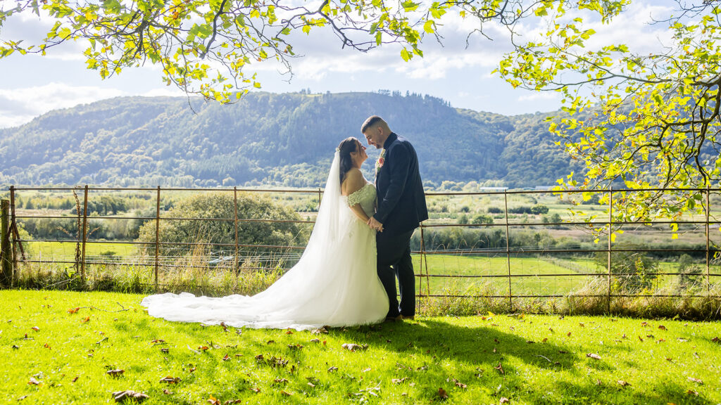 A bride and groom stand facing each other on a grassy lawn, with a wire fence and green hills in the background under a partly cloudy sky.