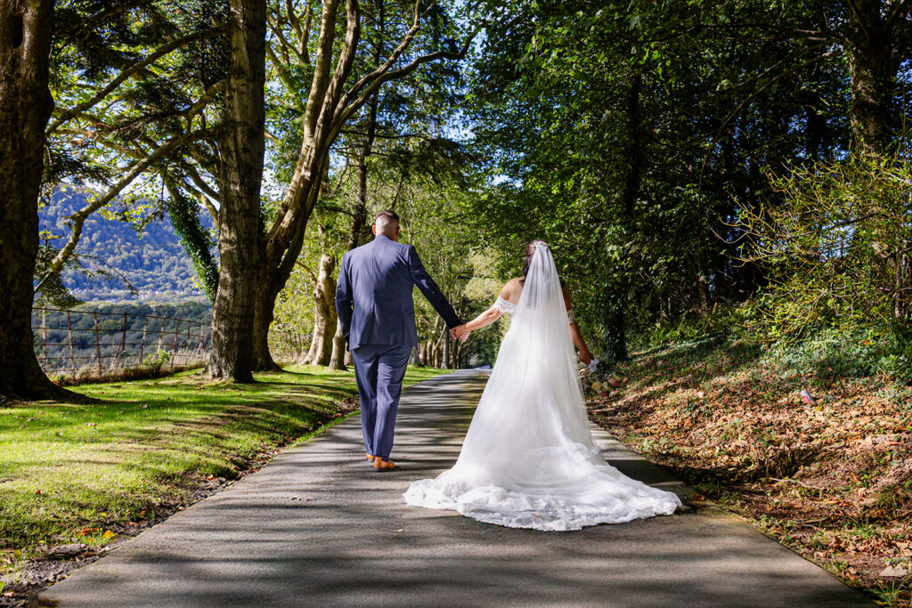 A bride and groom walk hand in hand down a sunlit, tree-lined path, with the bride in a white dress and veil, and the groom in a blue suit.