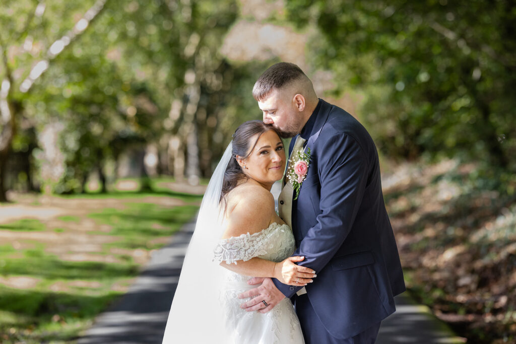 A bride and groom stand on a path in a park; the groom hugs and kisses the bride's forehead whilst she smiles at the camera.
