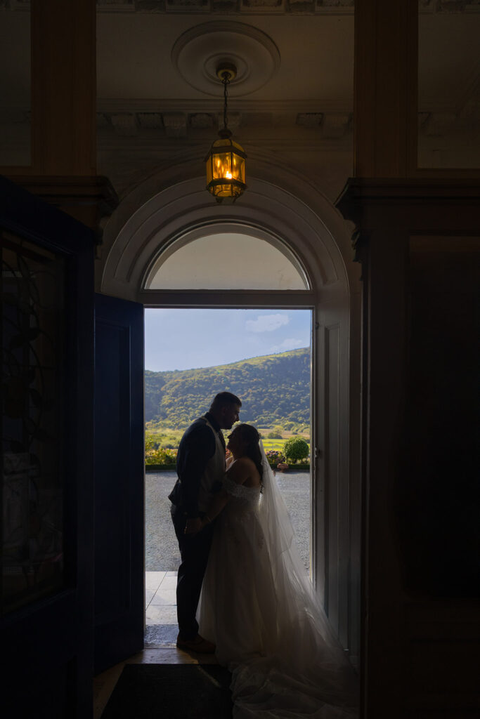 A bride and groom stand in the doorway of a building, silhouetted against natural light with mountains visible in the background.