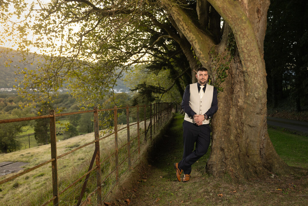 A man in formal attire leans against a large tree near a rusty fence, with grassy hills and trees in the background under an overcast sky.