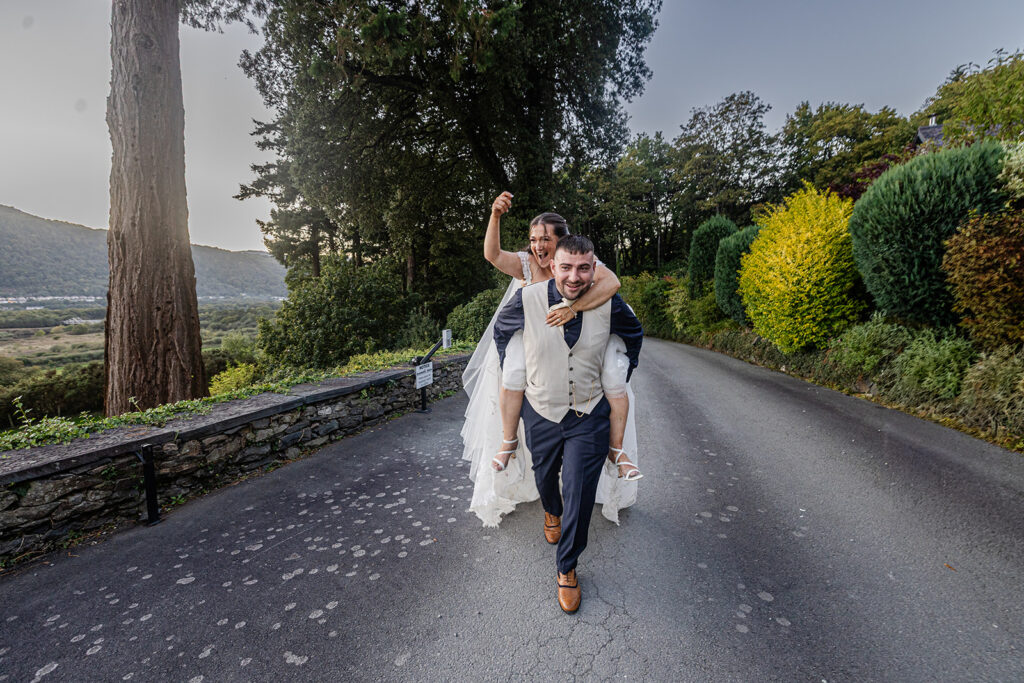 A bride in a white dress rides on the groom's back as they walk down a paved road surrounded by trees and greenery.