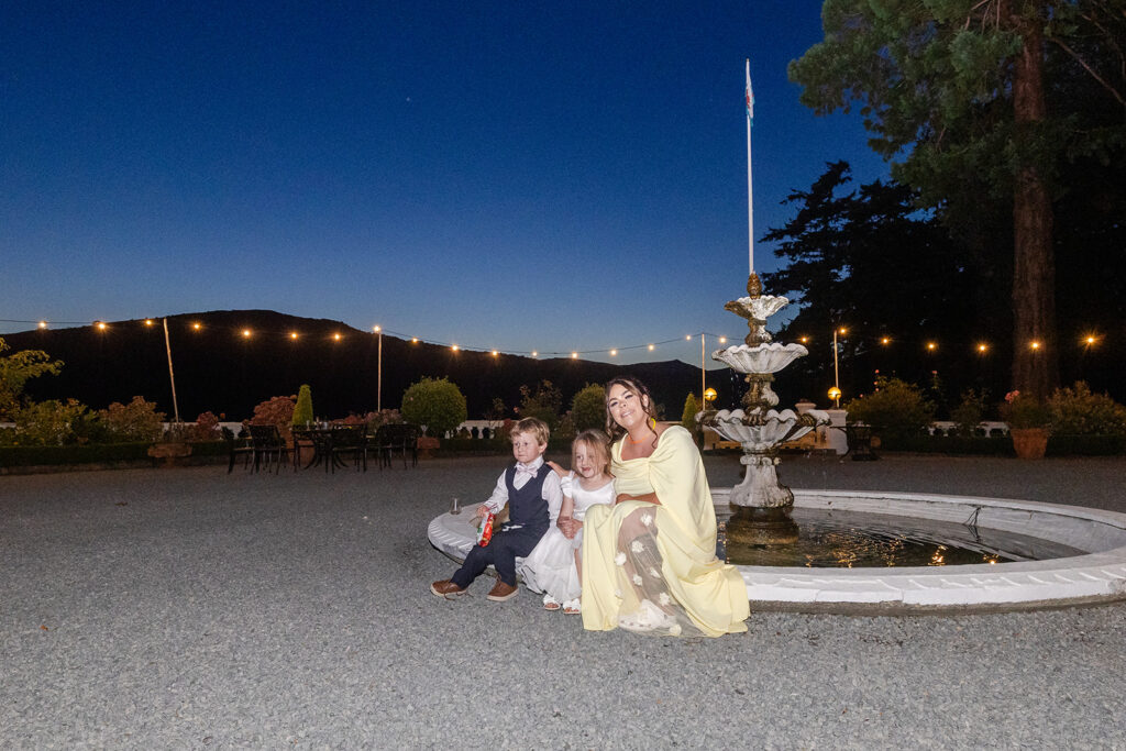 A woman and two children sit by a fountain in an outdoor area at dusk, with fairy lights and mountains visible in the background.