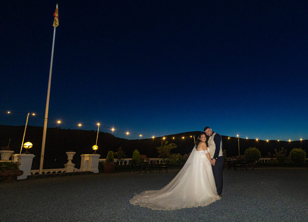 A bride and groom stand together outdoors at dusk, with fairy lights, a flagpole, and a dark blue sky in the background.