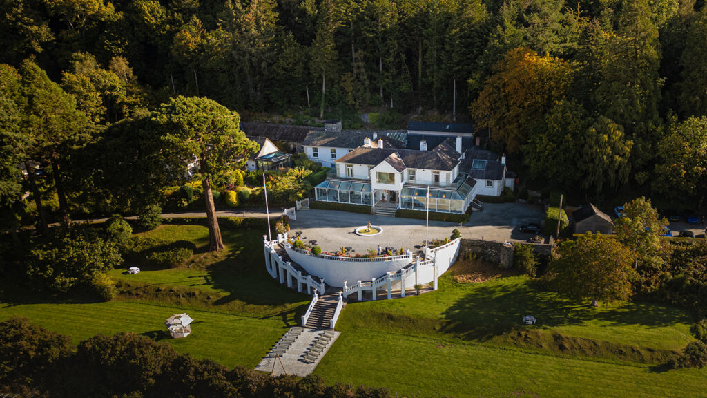Aerial view of a large white country house with a circular drive, surrounded by trees and landscaped gardens, with a lawn and steps leading to a lower terrace.
