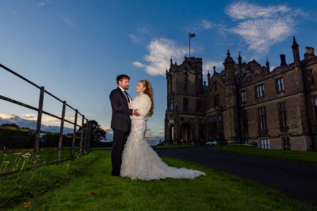 A bride and groom stand facing each other on grass near a large castle-like building under a blue sky with clouds.
